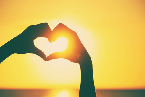 Hands forming the shape of a heart and framing a sunset or sunrise