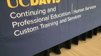 UC Davis logo on navy table banner with white text, wood floor beneath