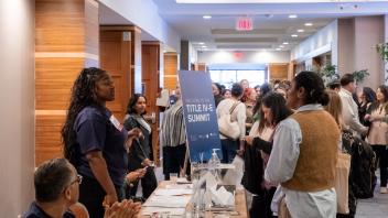 2025 Title Four E Summit participants gather at a registration table where Summit student volunteers wearing dark navy t-shirts are helping to sign people into the event.