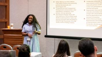 A young woman with dark hair wearing a blue and white dress speaks to a group of Title Four E Summit attendees. She stands near a lectern and is gesturing toward a projection screen with a slide displayed on it.