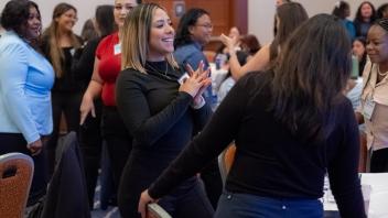 Two Title Four E Summit attendees laugh and chat in a busy ballroom during a break between presentations. They are both young women and they are both wearing dark tops.