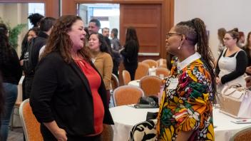 Two Title Four E Summit participants chat and laugh together during a break between presentations. The woman on the left is wearing a black jacket over a bright red shirt, and the woman on the right is wearing a long, colorful coat.