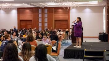 Avni Panchal, a Title Four E Summit presenter, stands on a raised stage at the front of a crowded banquet room, addressing Title Four E Summit participants. She is wearing a blue and red patterned dress and speaks into a hand-held microphone.