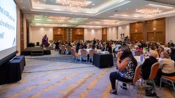 During a presentation by Title Four E Summit presenter Avni Panchal, Summit participants gather in a large banquet hall and watch the presentation. There is a large projection screen in the foreground, displaying a slide for Panchal's presentation.