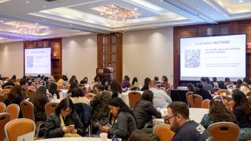 Lucy Fuentes, Title Four E Project Coordinator, stands at a lectern on a raised stage at the front of a crowded banquet hall during the Title Four E Summit. Participants sit around round banquet tables as Fuentes delivers her closing remarks. She is wearing glasses and a light-colored, floral-print dress.