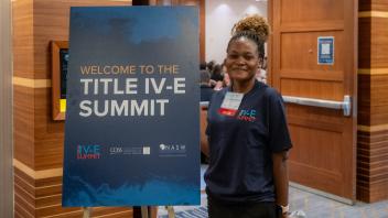 A Title Four E Summit student organizer wearing a navy blue staff t-shirt stands next to a large sign that reads 'Welcome to the Title Four E Summit.' She is smiling for the camera.