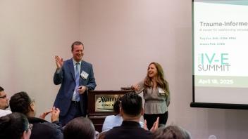 Two Title Four E Summit presenters, Tory Cox and Jessica Polk, stand at the front of a seminar room next to a lectern. They are smiling and speaking to a room full of Summit participants. Cox is wearing a suit and tie while Polk is wearing a floral patterned blouse.
