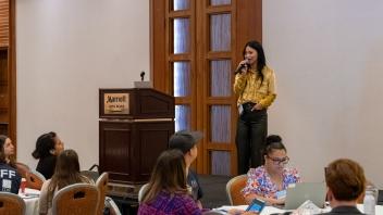 A Title Four E presenter, Christine Mark-Griffin, addresses a room full of Summit participants from a raised stage. She is standing next to a lectern and is wearing a bright yellow blouse and dark trousers.