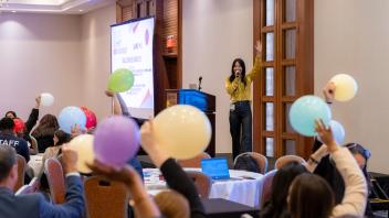 A Title Four E presenter, Christine Mark-Griffin, addresses a room full of Summit participants from a raised stage. She is standing next to a lectern and is wearing a bright yellow blouse and dark trousers. In the foreground, Summit participants hold up colorful balloons as part of an exercise that Mark-Griffin is leading from stage.
