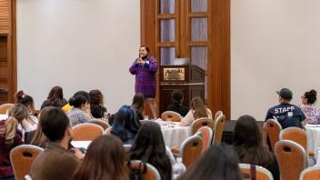 Avni Panchal, a Title Four E Summit presenter, stands on a raised stage at the front of a crowded banquet room, addressing Title Four E Summit participants. She is wearing a blue and red patterned dress and speaks into a hand-held microphone.