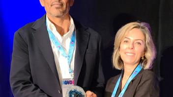 Two adults on stage smiling, holding a glass award with blue lanyards
