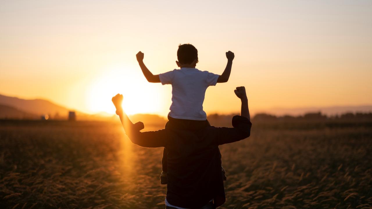 A grownup with a child on their shoulders. Both face the sun and pump their arms up in victory.
