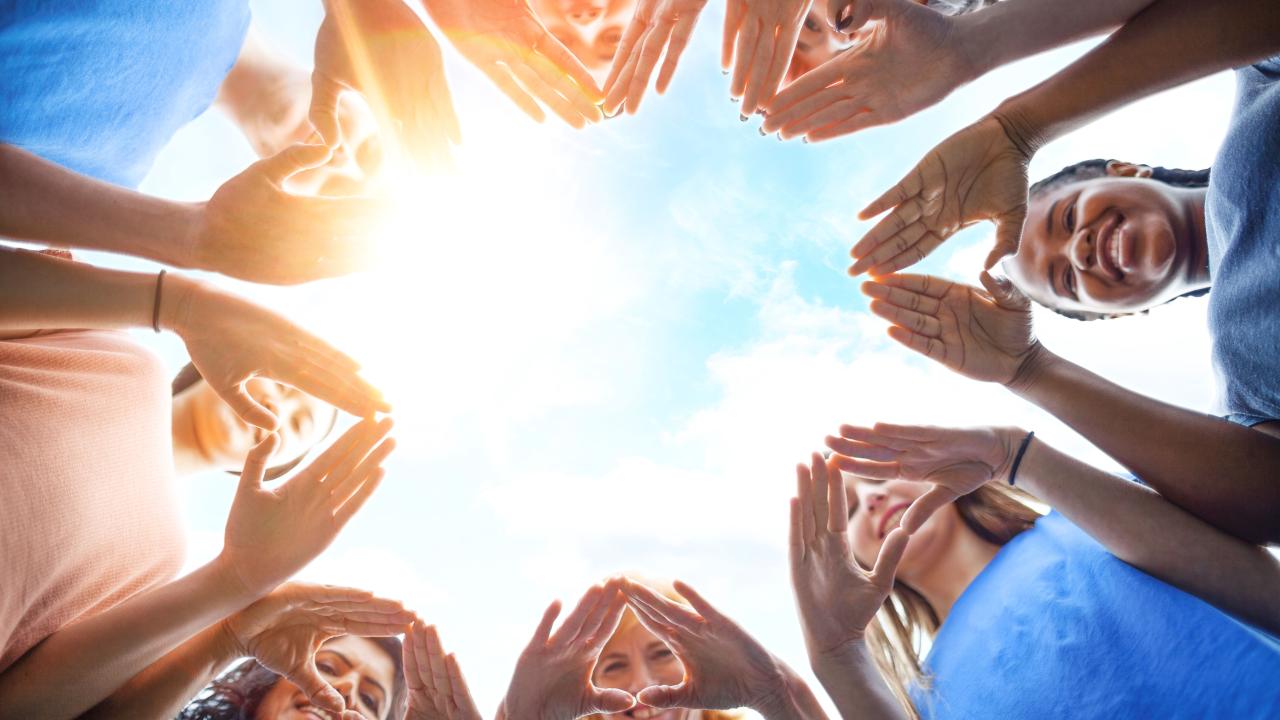 A circle of smiling people looking down at the camera, making heart shapes out of their hands.