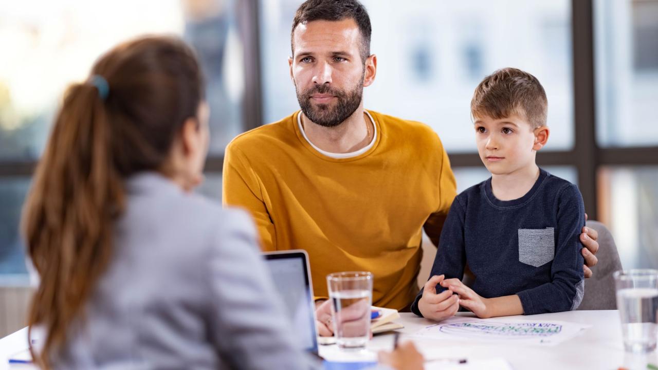 Man in yellow sweater and young son speaking with a woman across a desk in a bright office