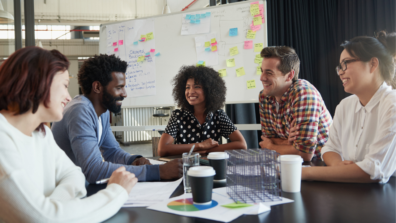 A group of coworkers gather around a table in a conference room, reviewing charts and graphs, suggesting that they are working together on a project. There is a whiteboard behind them covered with diagrams and post-it notes.