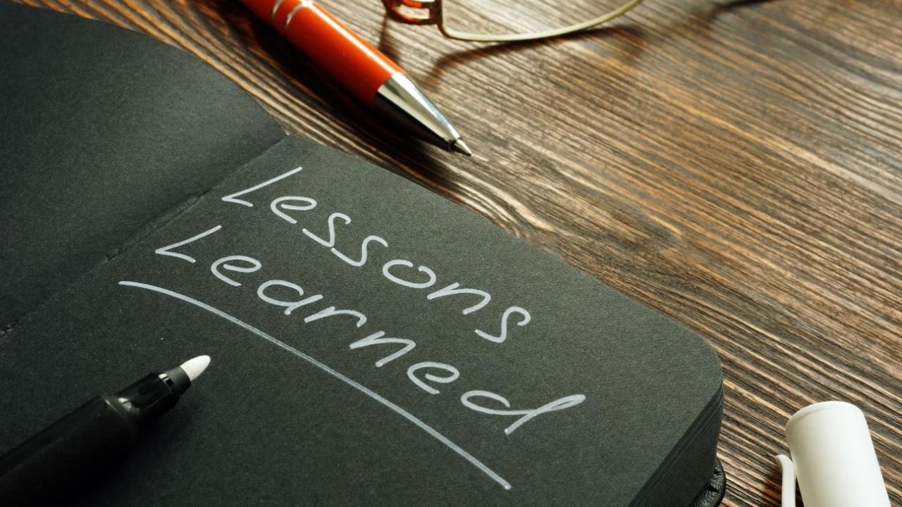 Black notebook with "Lessons learned" in white, pens and glasses on wooden desk