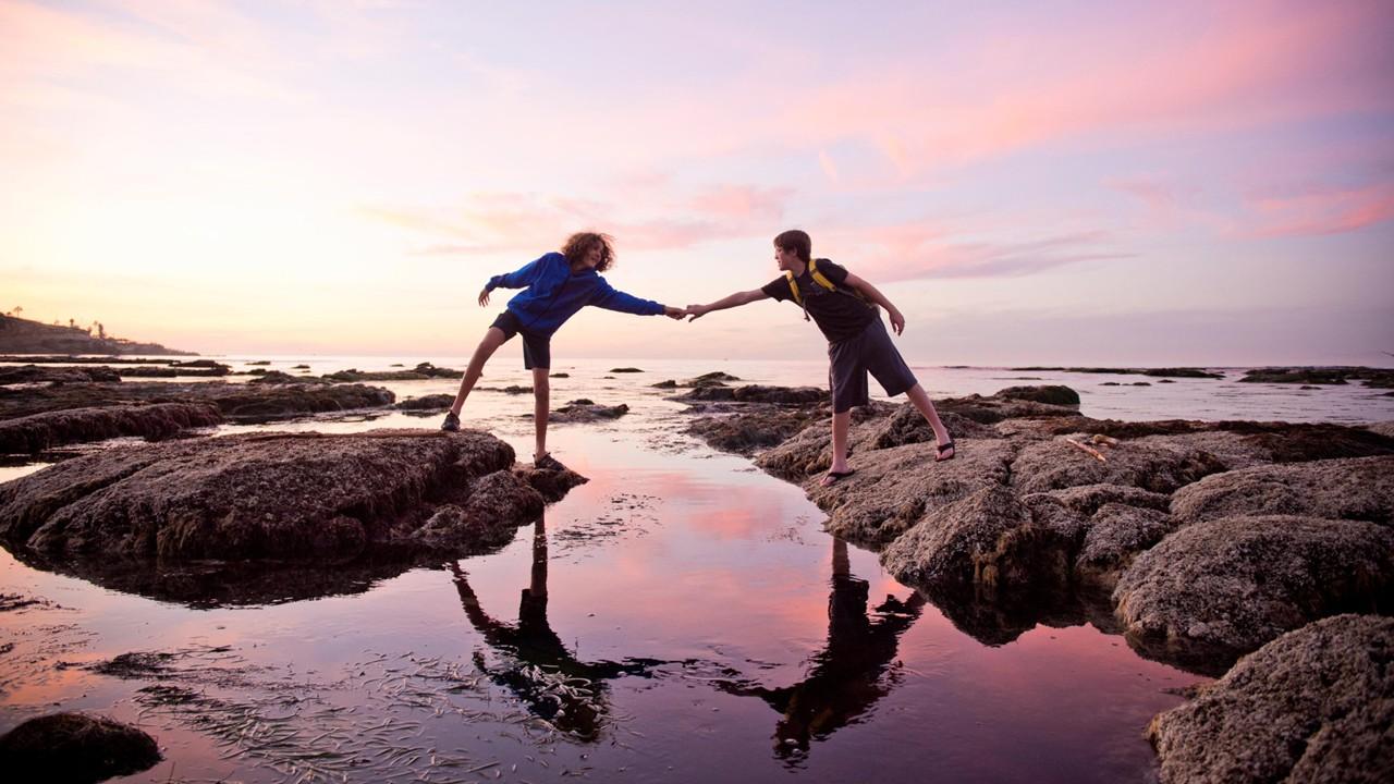 One hiker offers his hand to assist another hiker across a stream.