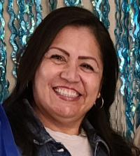 Portrait photo — smiling woman with dark hair, hoop earrings, denim jacket, blue streamer backdrop