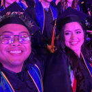 Three smiling graduates in caps and gowns under purple stage lighting