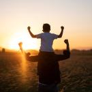 A grownup with a child on their shoulders. Both face the sun and pump their arms up in victory.