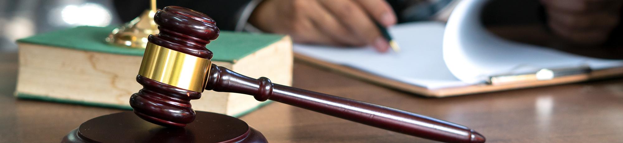 gavel on desk in foreground with law book and man taking notes in background